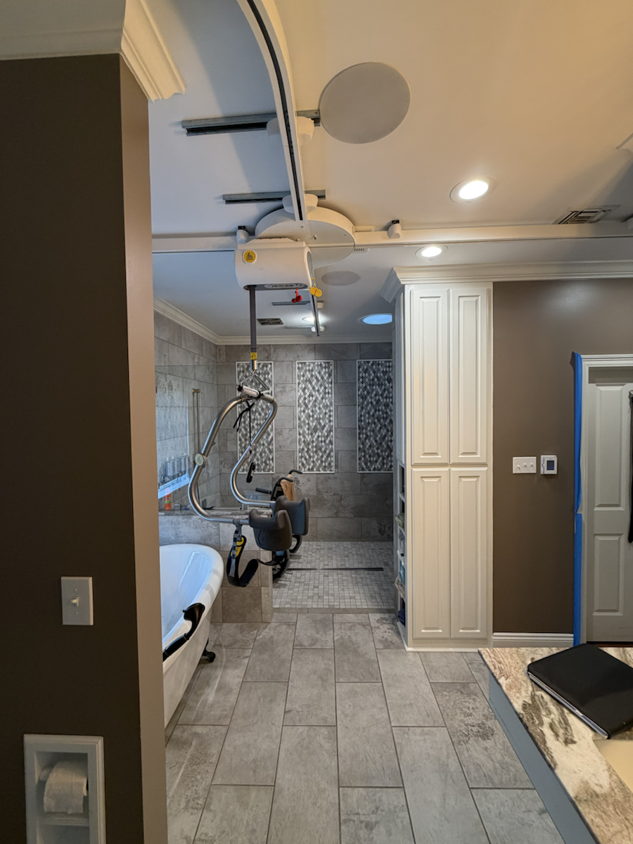 Ceiling lift rail system installed in a bathroom for assisted transfers near a bathtub and shower.
